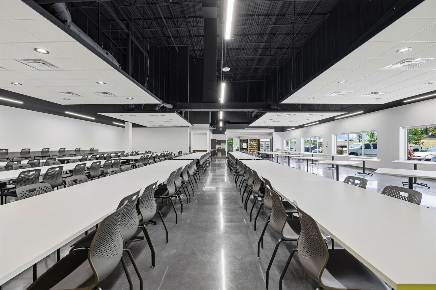 Large staff cafeteria with long white tables, black chairs and a polished concrete floor