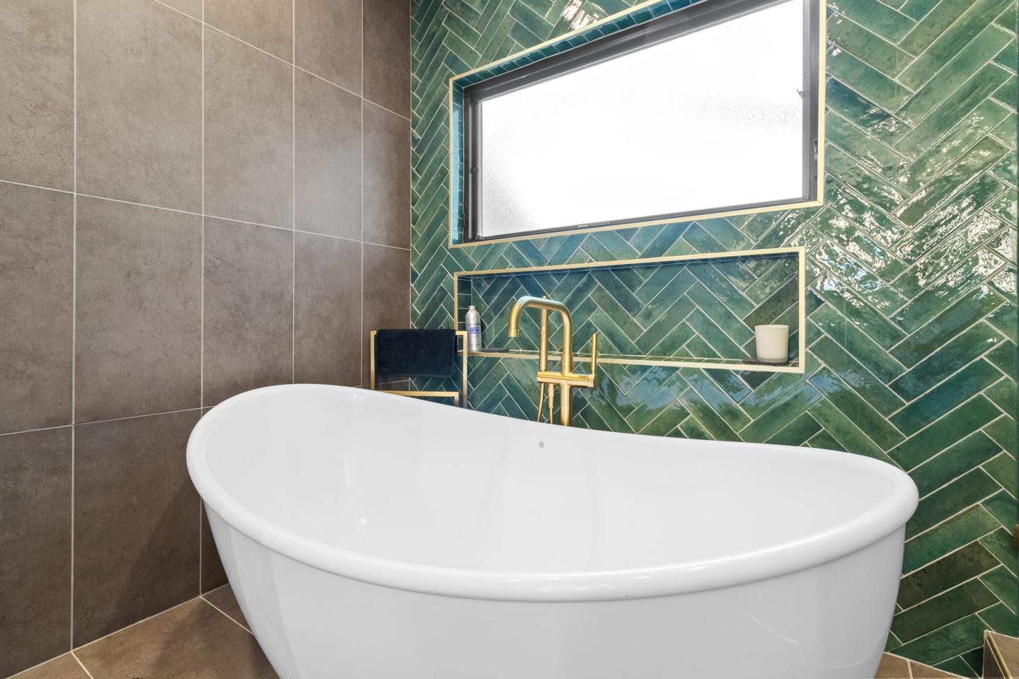 Freestanding soaking tub against a green tiled wall with a gold filler and a picture window