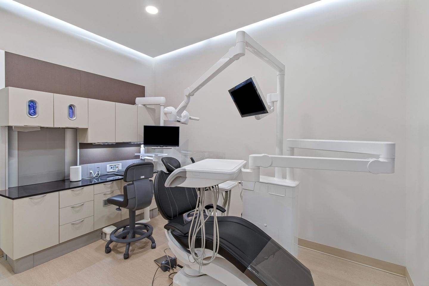 Dental operatory with a black patient chair, an overhead monitor arm and soft cove lighting