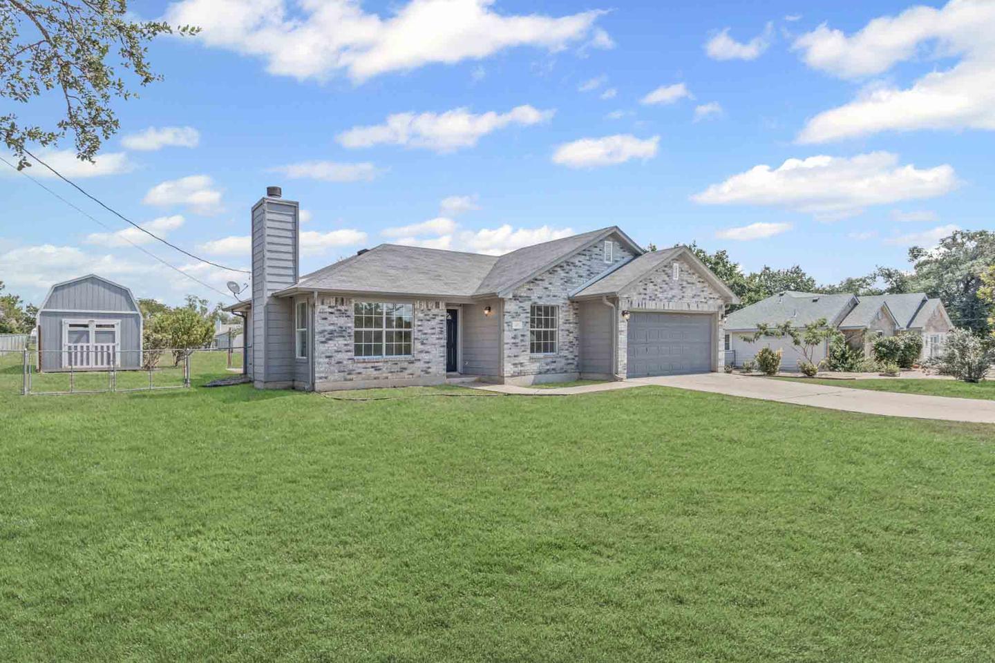 Single story stone home in Liberty Hill across a wide mown lawn, with a gray storage barn beside it and a neighboring house beyond