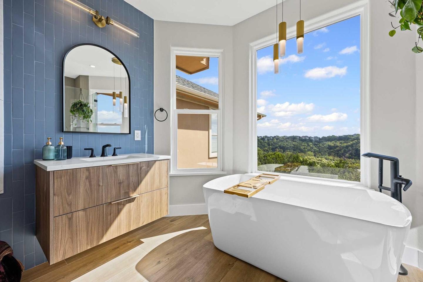 Primary bathroom with blue tile, a floating walnut vanity and a freestanding tub beside a window