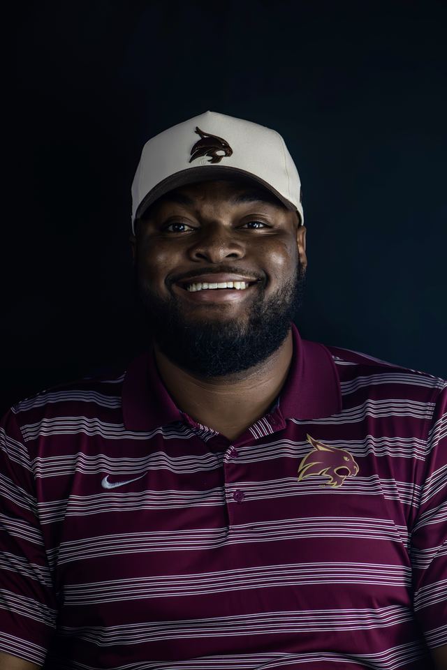Relaxed headshot of a smiling man in a baseball cap and striped polo against a black backdrop