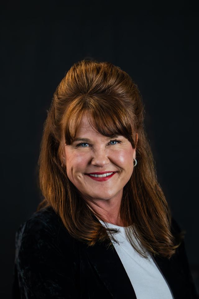 Headshot of a smiling woman with auburn hair in a black jacket against a black studio backdrop