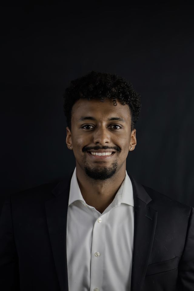 Headshot of a young man in a black suit and white open collar shirt against a black studio backdrop