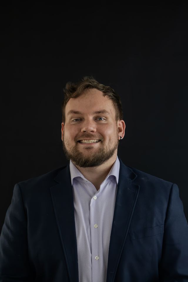 Headshot of a smiling man with a beard in a navy blazer and white shirt against a black studio backdrop